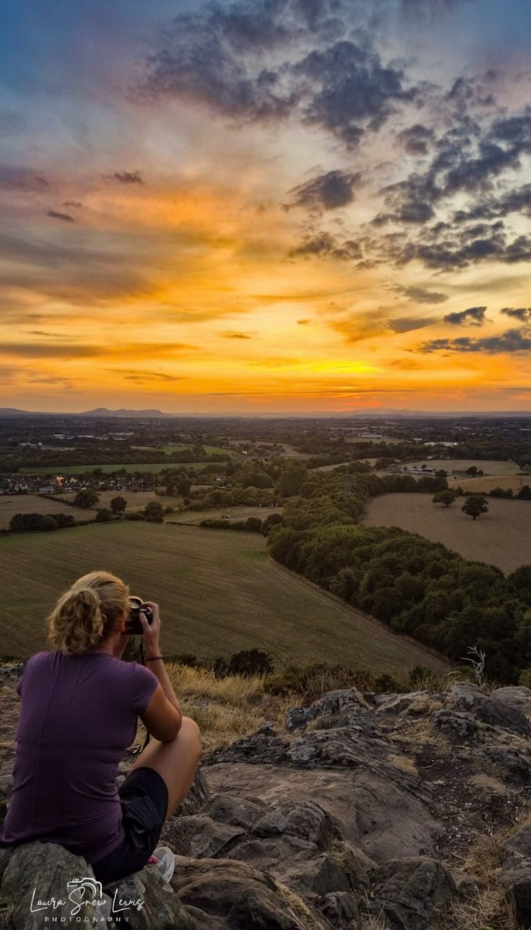 A person photographing a sunset over a serene landscape with fields and hills.