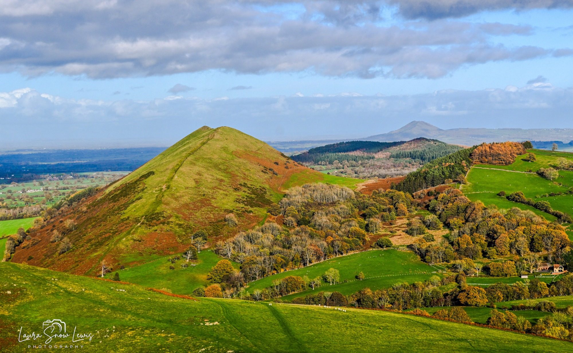 Rolling green hills, the Lawley under a partly cloudy sky, with the Wrekin in the distance.