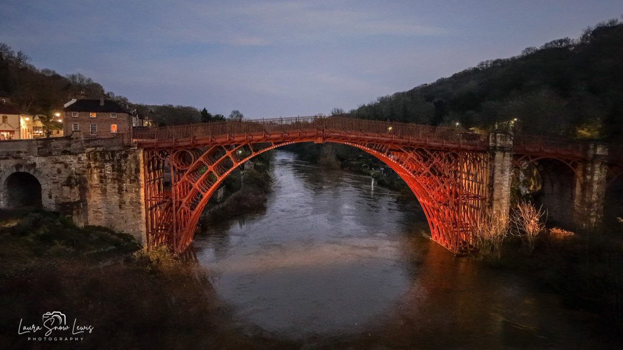 A red iron bridge spanning a tranquil river at dusk, surrounded by lush greenery. Ironbridge, Telford