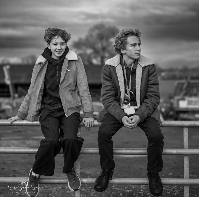 Two young adults sit on a fence, one smiling and the other looking away, in a cloudy setting.