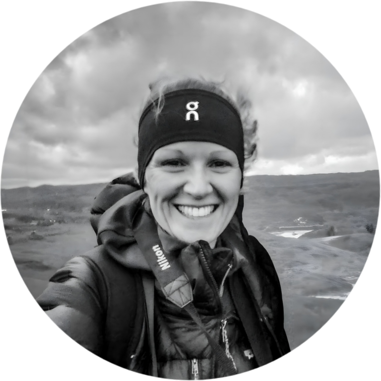 Smiling woman in outdoor gear against a scenic landscape with cloudy skies.