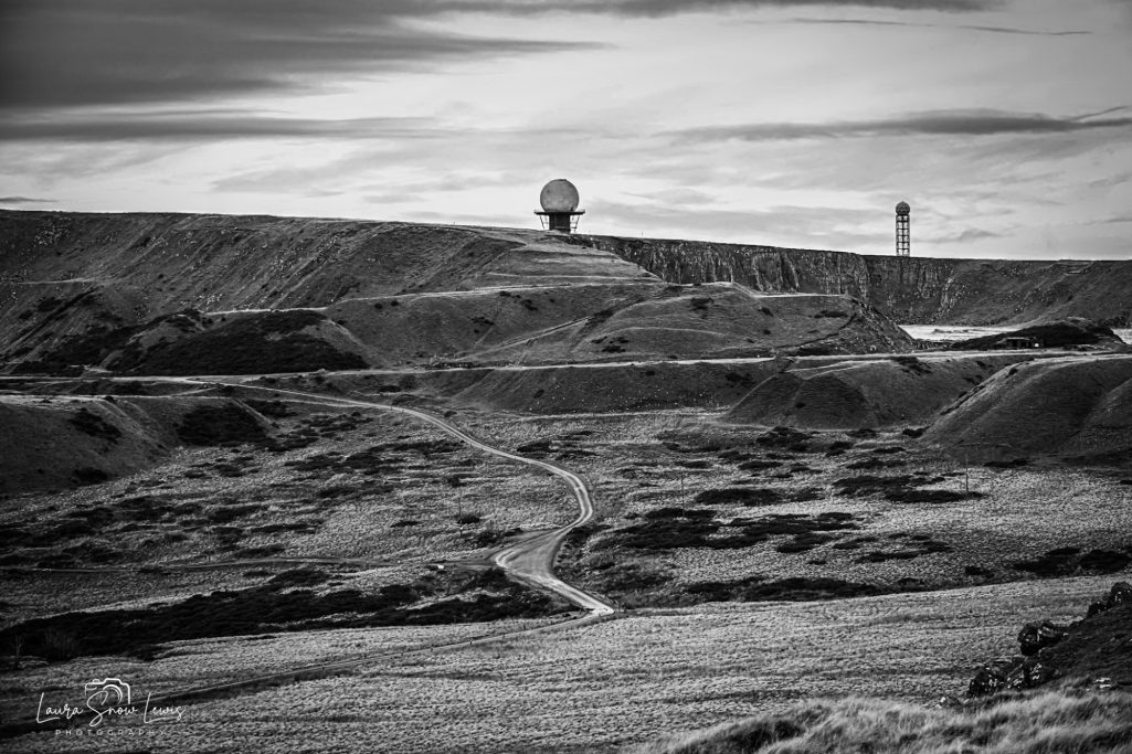 Desolate landscape with hills, two radar domes, and a winding path in black and white.